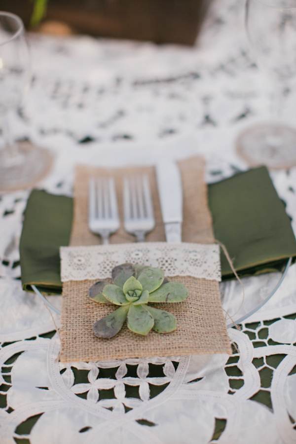 A place setting with two forks and a knife on a green napkin, topped with burlap, lace, and a small succulent, rests on a white lace tablecloth—perfect wedding inspiration for doily ideas.
