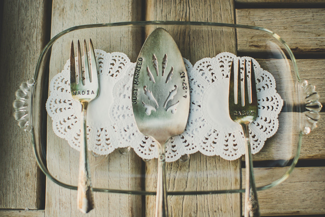 A glass dish with doily wedding inspiration: two doilies, forks labeled "NADIA" and "CASEY," and a pie server engraved with "JUST MARRIED 4.27.15," all arranged on a wooden surface.
