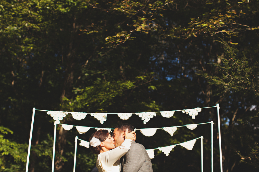 A couple dressed in wedding attire kisses outdoors under lace bunting, offering charming doily wedding inspiration with trees and sunlight in the background.
