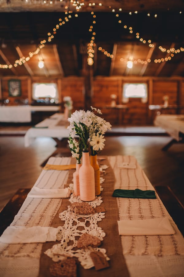 A rustic table is decorated with lace runners, crochet doilies perfect for a doily wedding, pastel bottles holding white flowers, and surrounded by string lights in a wooden room—charming wedding inspiration.