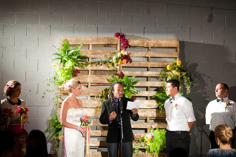 Couple with officiant at ceremony in front of a rustic wood pallet backdrop layered with colorful tropical and garden flowers and greenery