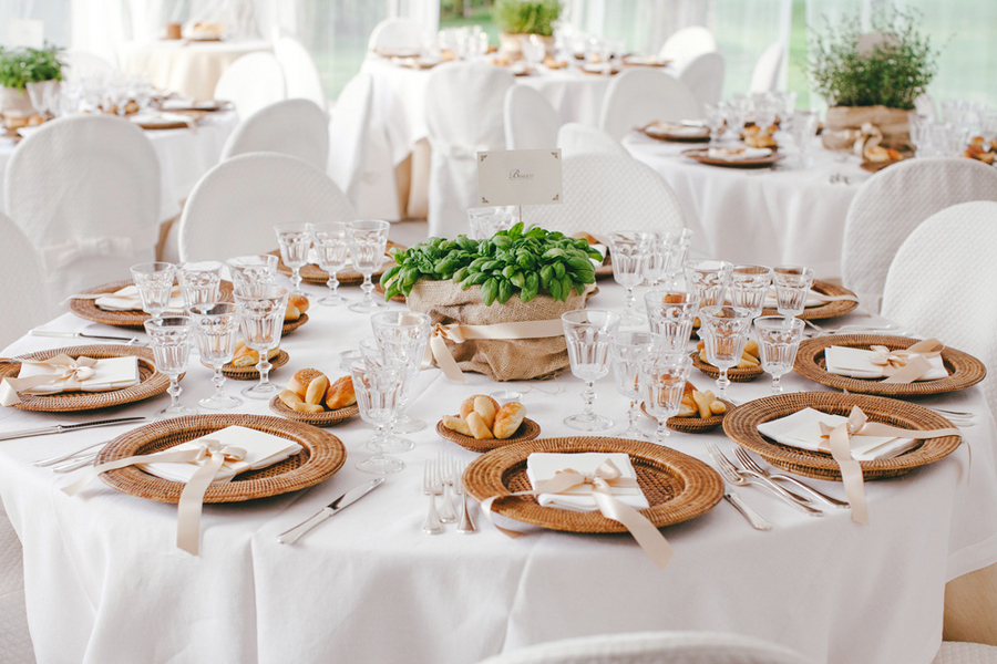 Simple round wedding table with white linen, woven rattan chargers, white napkins, and a potted basil plant wrapped in burlap as the centerpiece