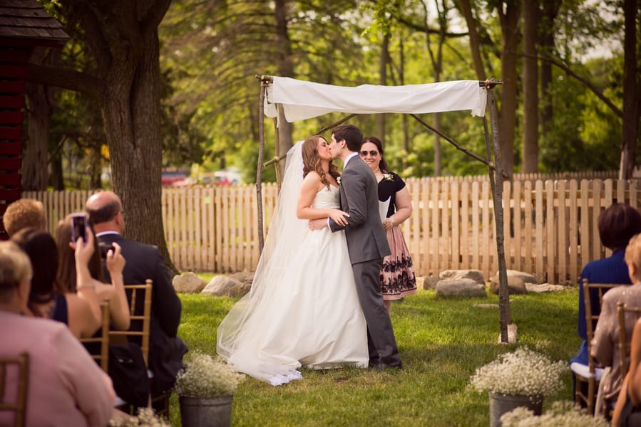 Bride and groom kissing under an outdoor wooden frame ceremony canopy with white draped fabric panels in a garden wedding setting