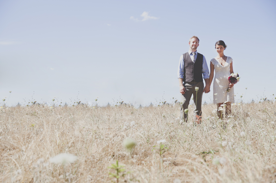 Bride and groom standing in wheat field under blue sky, bride in ivory lace tea-length dress with cap sleeves