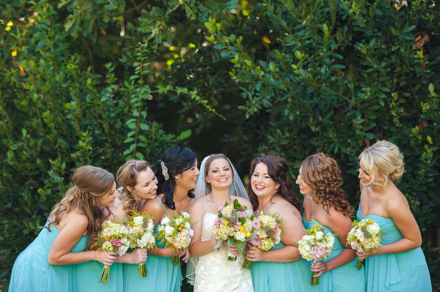 Five bridesmaids in matching tiffany blue strapless floor-length chiffon gowns laughing together against a lush green hedge backdrop with colorful wildflower bouquets at a California garden wedding