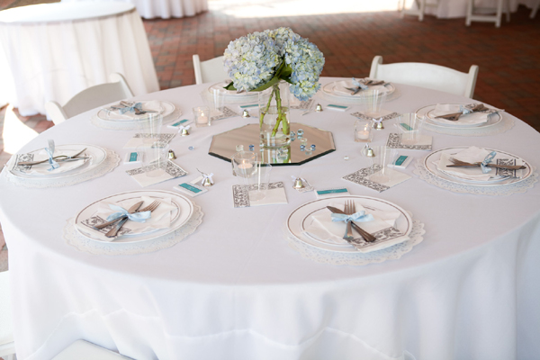 Round wedding reception table with blue hydrangea centerpiece in glass vase and elegant place settings with blue accents