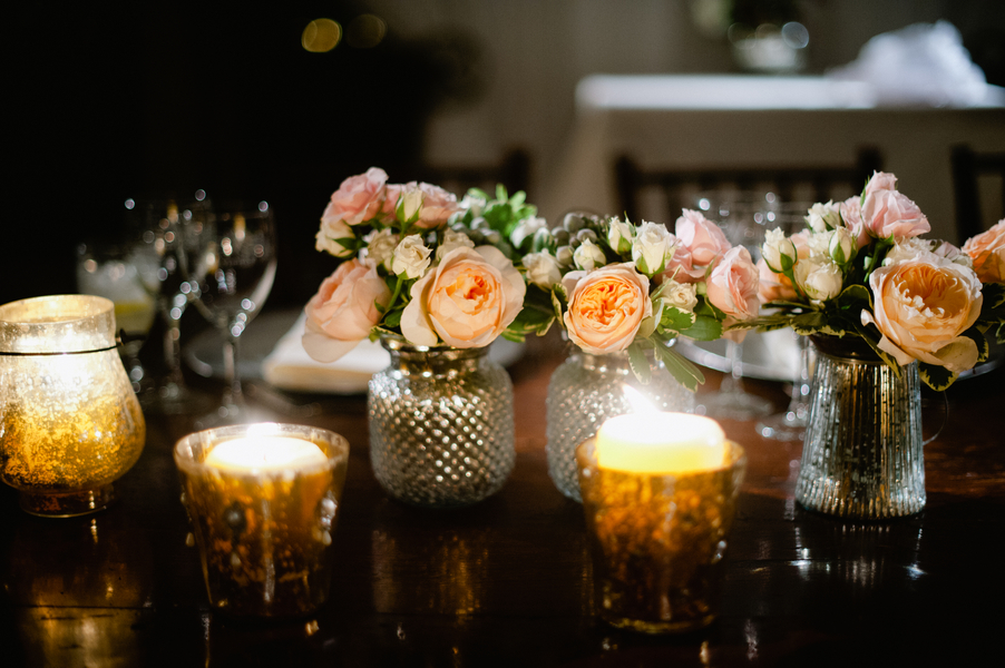 Low peach and blush rose arrangements in mercury glass and hobnail vases flanked by mercury glass votive candles on a dark wood table