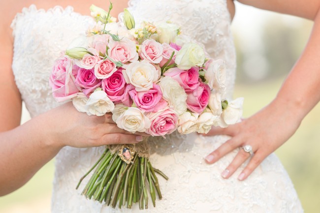 Bride holding rounded bouquet of pink and white spray roses with heirloom brooch on stem wrap