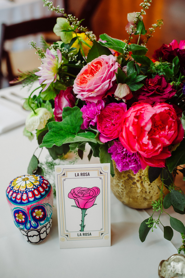 Vibrant Day of the Dead centerpiece with hot pink roses and coral peonies beside a painted sugar skull and Loteria card