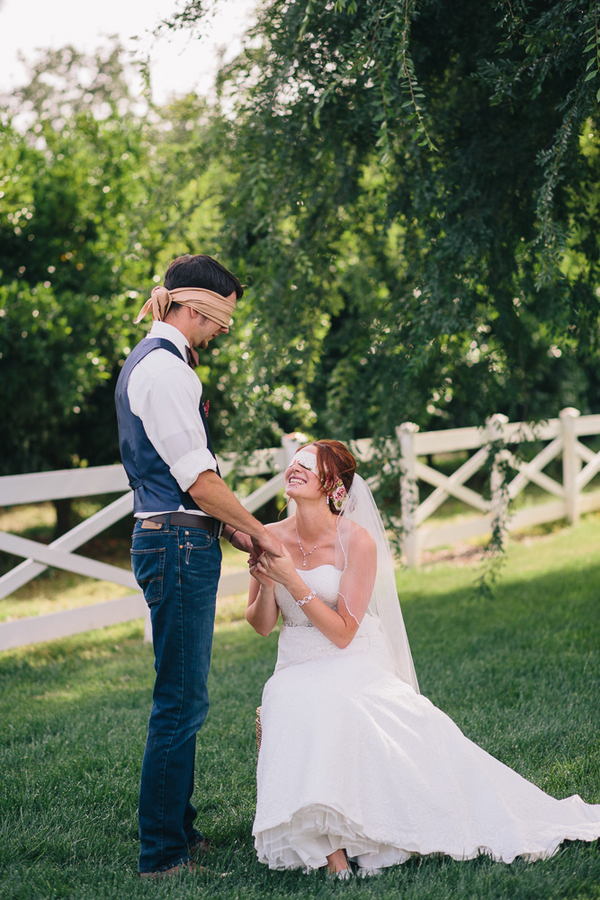 Bride kneeling before a blindfolded groom on a green lawn with a white post-and-rail fence and lush trees behind