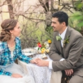 bride wearing blue plaid and groom sitting at a table with lace table runner and pink, yellow and green flowers