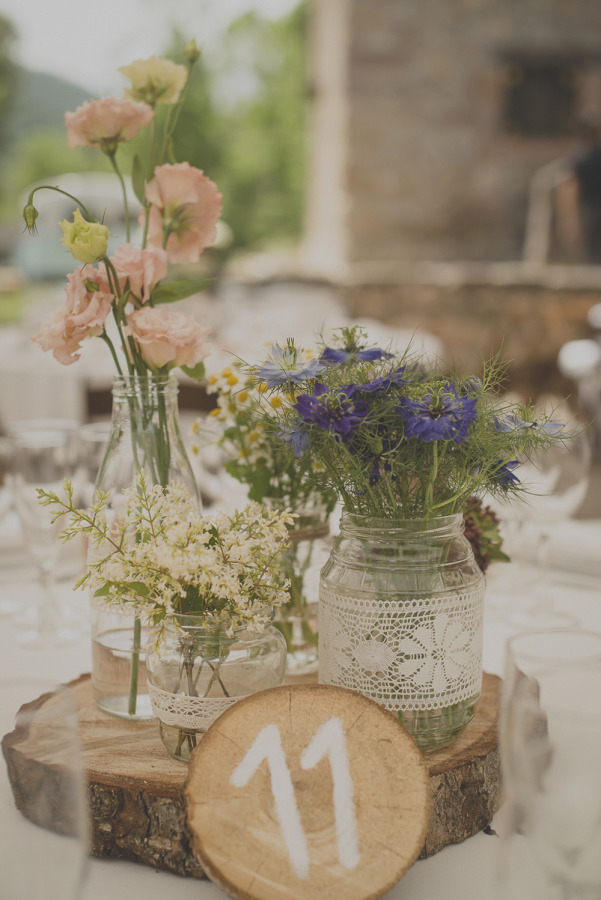 Assorted wildflowers in glass jars and bottles, some wrapped in lace, arranged on a wooden log slice with a table number