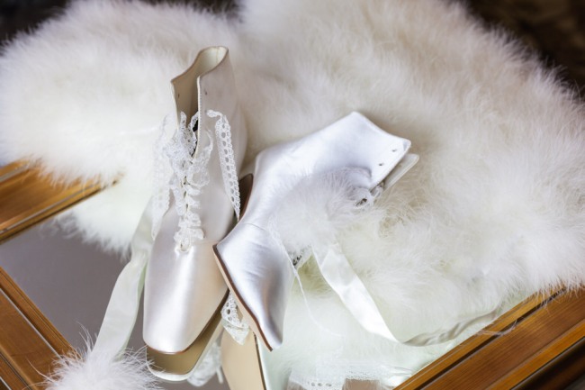 White satin Victorian lace-up bridal booties with eyelet trim resting on a white feather boa