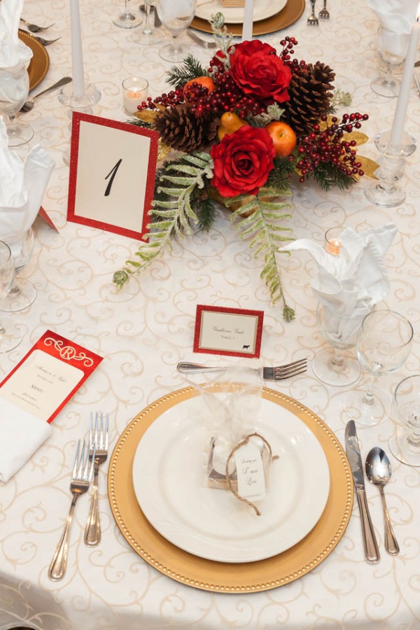 Winter wedding table with gold scroll linen, gold beaded chargers, and a centerpiece of red roses, pinecones, berries, and evergreen