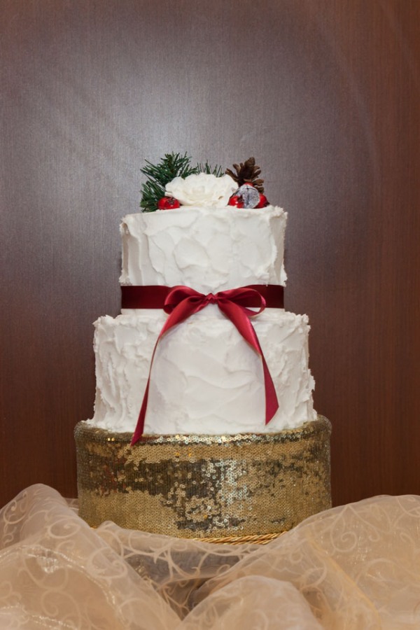 Two-tier snow-textured white buttercream wedding cake with a cranberry ribbon bow and a pinecone and berry topper on a gold sequin stand