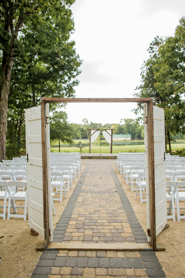 Freestanding white vintage doors framing a cobblestone aisle leading to a draped arch, with green trees on either side