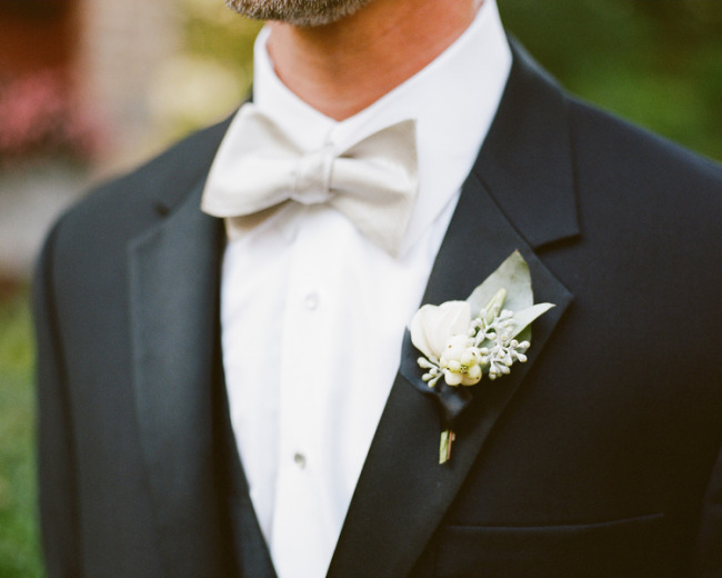 Small white bloom with sprays of tiny pale berries and one silvery grey-green leaf, pinned to a black tuxedo with an ivory bow tie