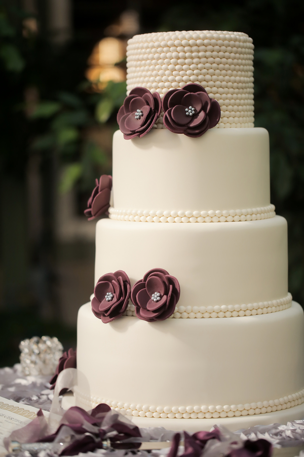 Four-tier white wedding cake featuring pearl-like accents and dark purple fondant flowers with jeweled centers, beautifully showcasing one of the latest wedding cake styles, displayed on a table with blurred greenery in the background.