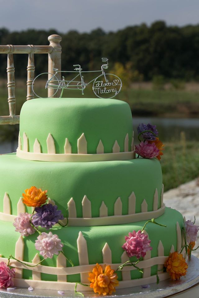 Three-tiered green cake in charming wedding cake styles, decorated with a white picket fence design, colorful flowers, and a wire topper shaped like a tandem bicycle with the names “Florian” and “Melany.”.