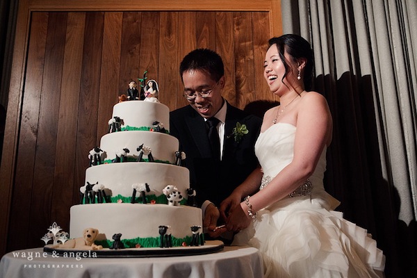 A bride and groom cut a tiered wedding cake decorated with dog figurines, standing together and smiling in formal attire—a charming example of unique wedding cake styles.
