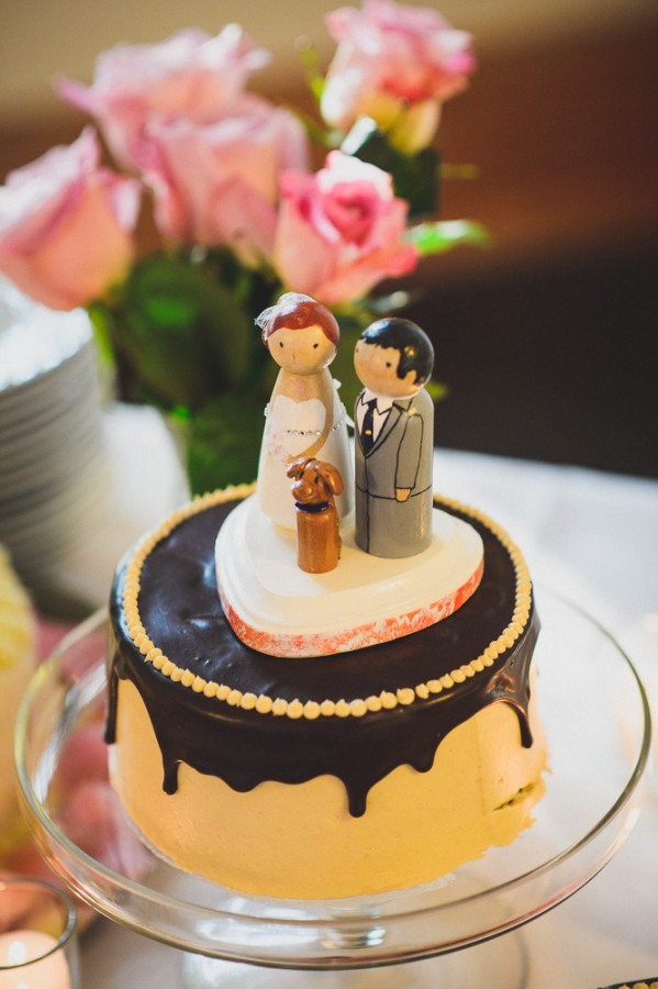 A round cake with chocolate drip icing showcases a bride and groom figurine and a small dog on top, with pink roses in the background—perfect for those seeking unique wedding cake styles.