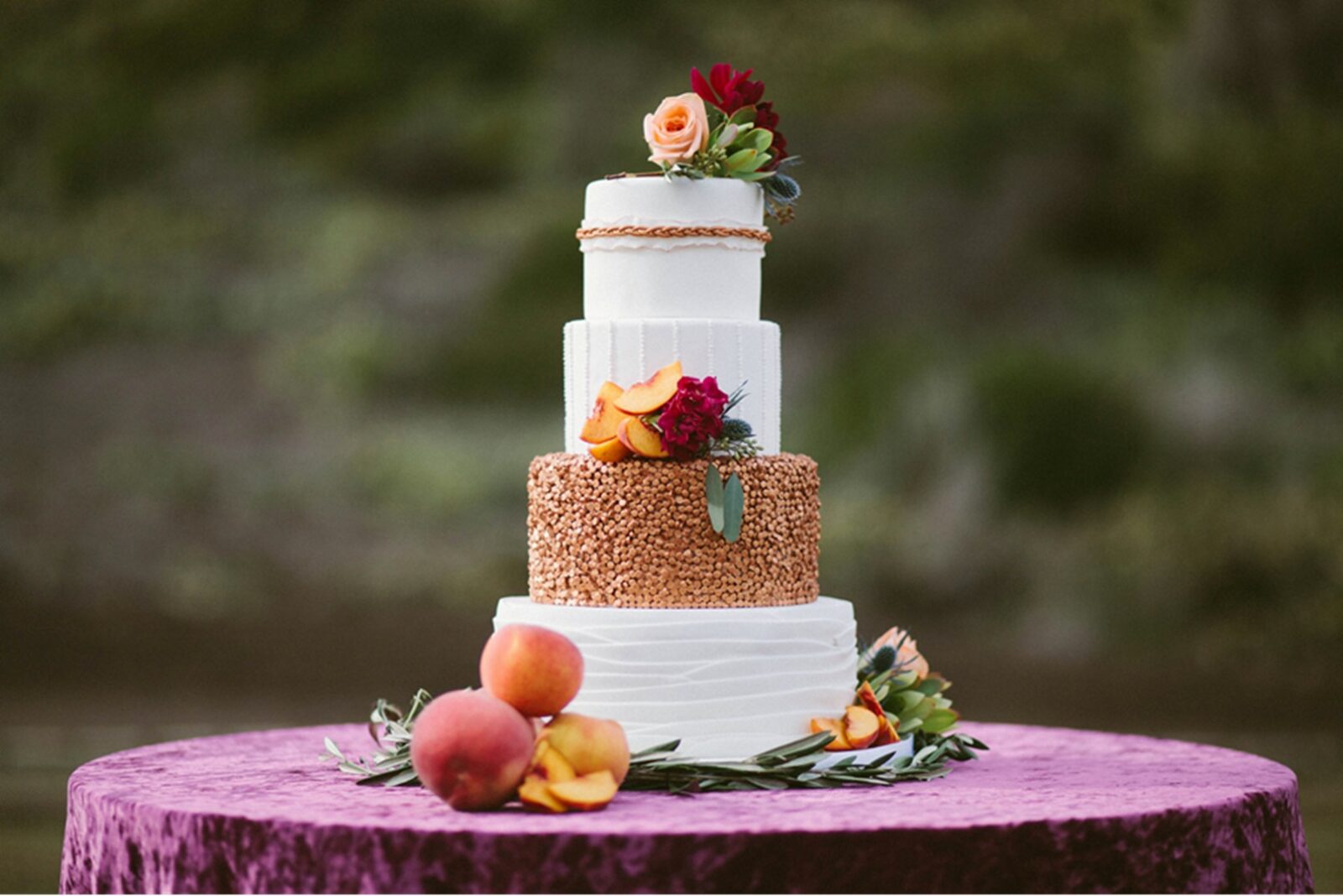 A three-tiered wedding cake showcasing elegant wedding cake styles, decorated with flowers and gold accents, is placed on a purple velvet tablecloth with peaches and greenery around the base.