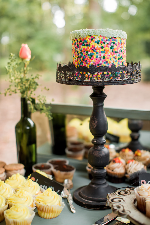 A multicolored confetti cake, inspired by playful wedding cake styles, sits on a black ornate stand surrounded by yellow cupcakes and other desserts, with a flower in a bottle in the background.