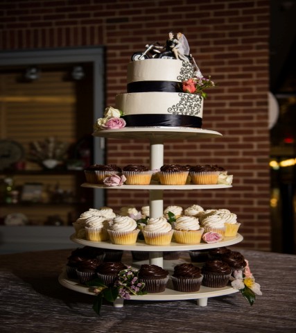 A tiered stand holds chocolate and vanilla cupcakes with a two-layer white cake on top, decorated with a bride and groom figurine and flowers—a charming display inspired by classic wedding cake styles.