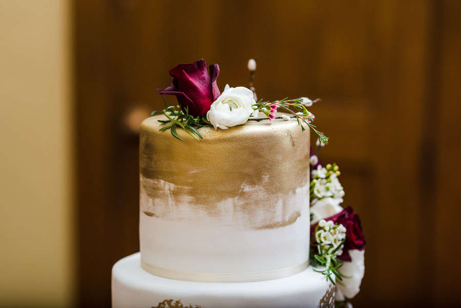 A two-tiered cake inspired by elegant wedding cake styles, with a gold-painted top tier, decorated with white and dark red flowers and green foliage, set against a brown background.