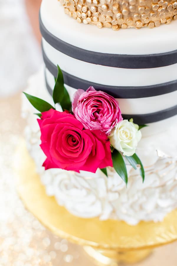A white tiered cake in elegant wedding cake styles, featuring black stripes, gold accents, and adorned with pink and white roses with green leaves on a gold cake stand.