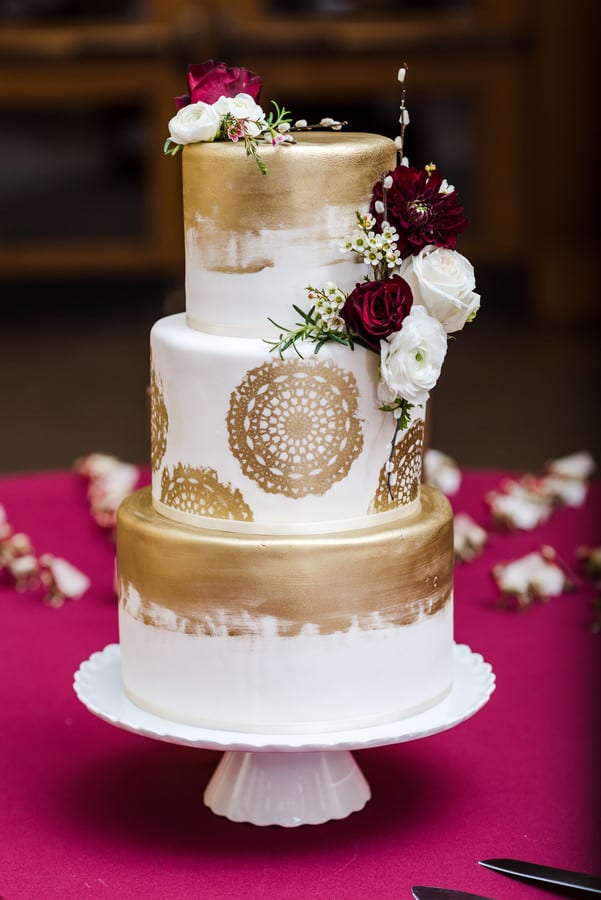 Three-tiered white cake with gold accents and lace patterns, showcasing popular wedding cake styles, decorated with red and white flowers on a white cake stand against a burgundy tablecloth.