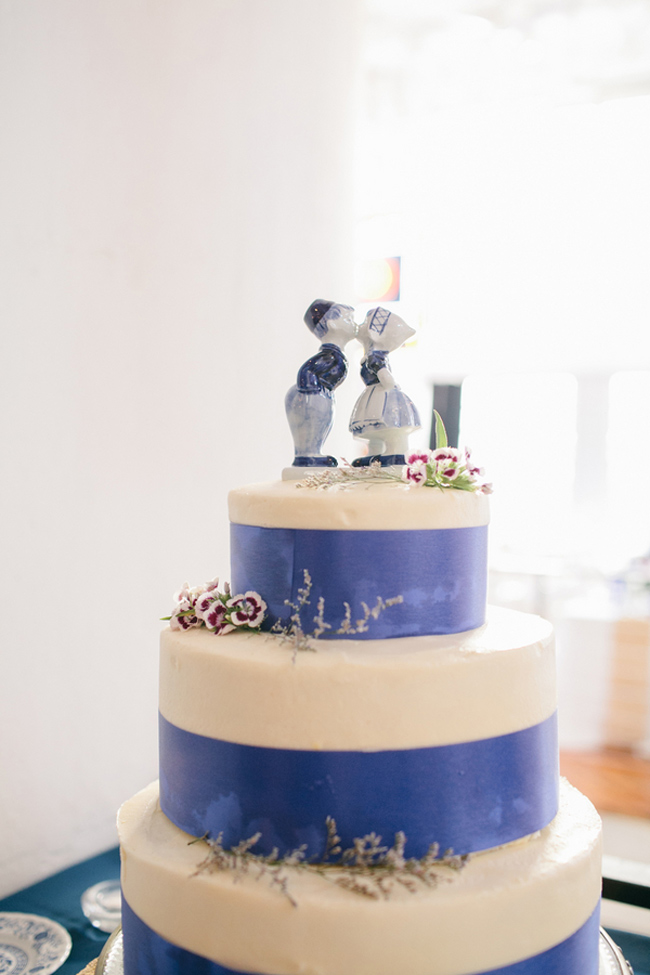 Three-tiered white cake with blue ribbon accents, decorated with purple flowers and topped with ceramic figurines of a kissing couple in traditional Dutch attire—a charming nod to classic wedding cake styles.
