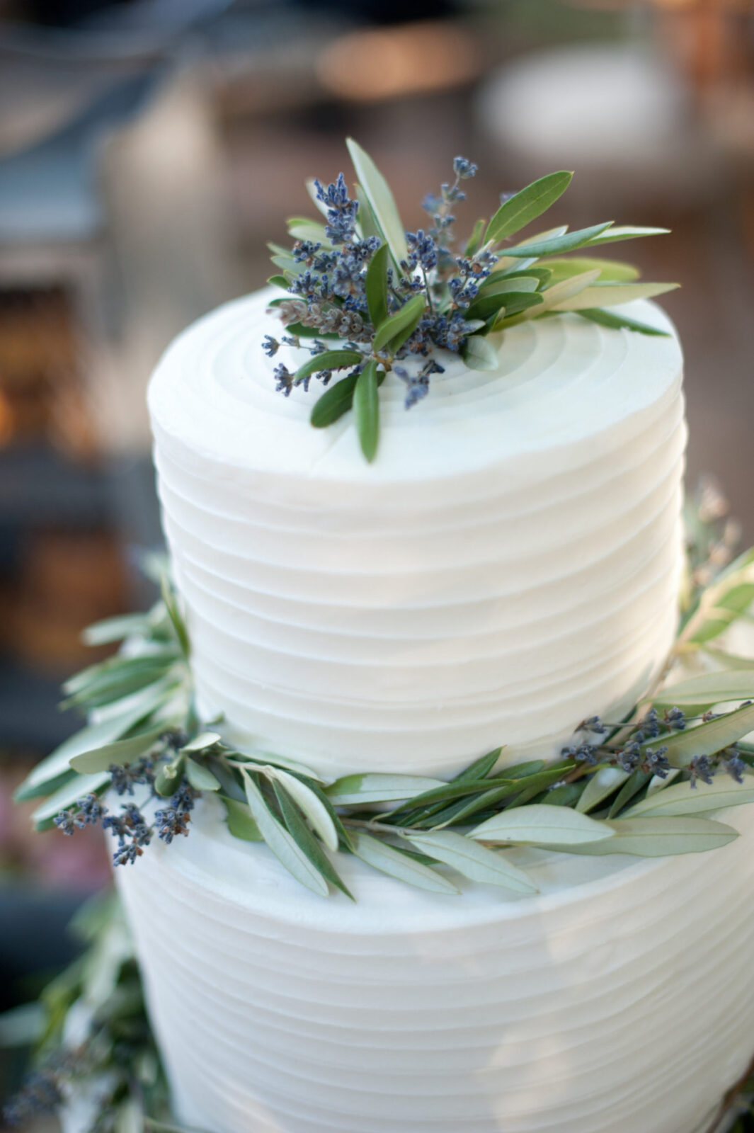 A white, two-tiered cake decorated with green leaves and small purple flowers, featuring textured horizontal lines on the icing—an elegant choice inspired by popular wedding cake styles.