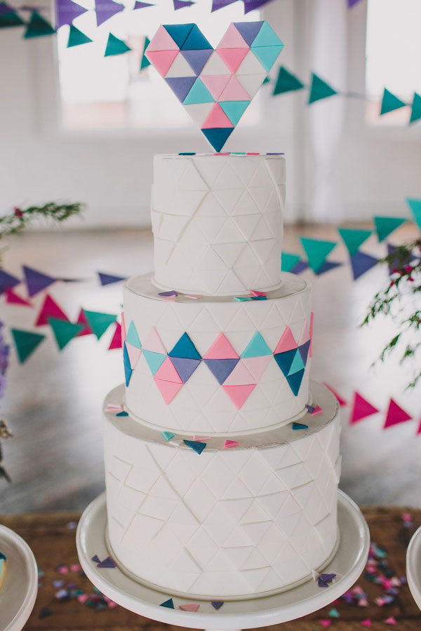 A three-tiered white cake, inspired by modern wedding cake styles, is decorated with geometric pastel triangles and topped with a heart-shaped arrangement of colorful triangles. Triangular bunting hangs in the background.