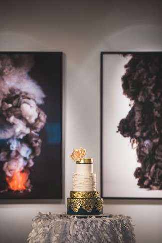 A tiered white and gold wedding cake, showcasing elegant wedding cake styles with a flower topper, is displayed on a round table in front of two abstract art pieces featuring smoke-like forms.