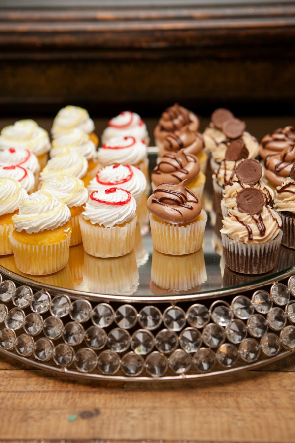 A tray with three rows of decorated cupcakes showcases wedding cake styles: vanilla with white frosting, vanilla with white frosting and red swirls, and vanilla with chocolate frosting and chocolate toppings.
