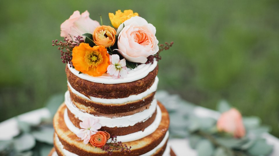 A three-tiered naked cake with white frosting between layers, showcasing popular wedding cake styles, is decorated with orange, pink, and yellow flowers on top and set outdoors.