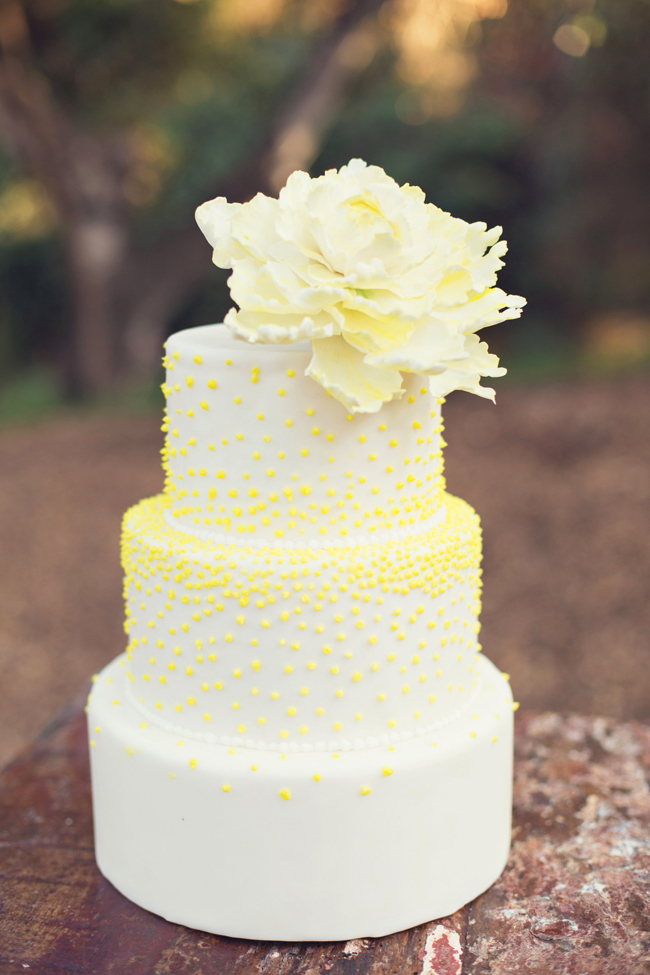 This three-tier white cake, decorated with small yellow dots and topped with a large white flower, perfectly showcases elegant wedding cake styles on a rustic wooden surface outdoors.