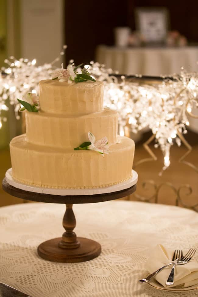 Three-tiered frosted cake featuring wedding cake styles, adorned with white flowers and green leaves on a wooden stand, set atop a lace-covered table and surrounded by glowing string lights.