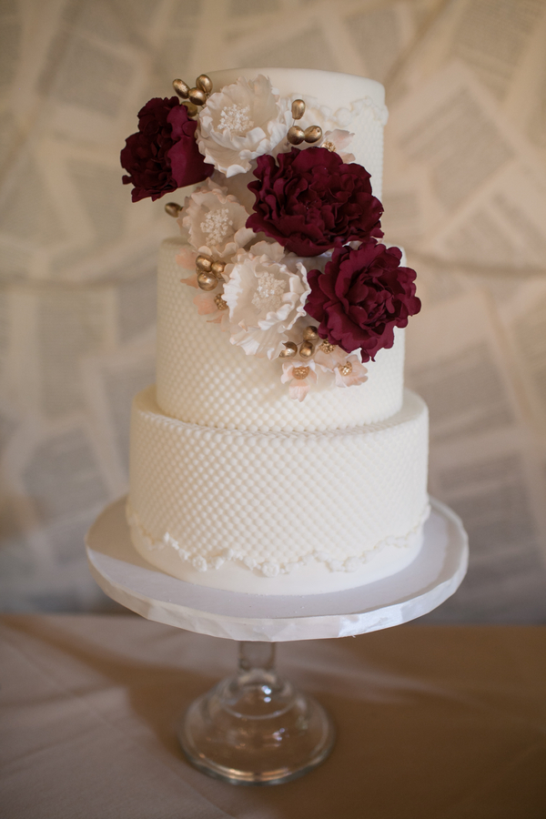 A three-tiered white wedding cake with textured icing, showcasing popular wedding cake styles, decorated with burgundy and white flowers and gold accents, displayed on a glass cake stand.