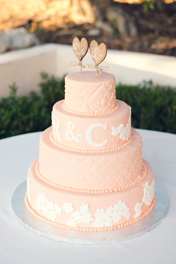 Three-tiered pink wedding cake with white floral decorations, bead borders, initials "R & C," and two heart-shaped toppers reading "We Do," beautifully showcasing one of the most romantic wedding cake styles outdoors on a white table.