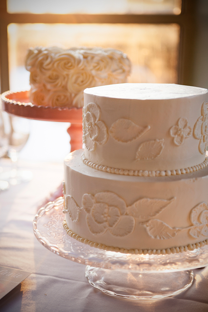 Two white cakes showcasing elegant wedding cake styles—one two-tiered with floral icing designs and one single-tiered with rosette frosting—are beautifully displayed on glass stands near a sunlit window.
