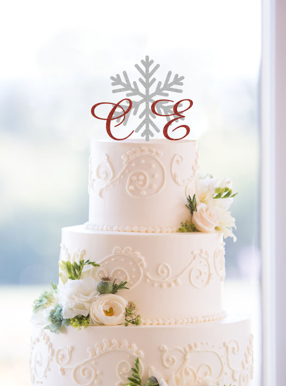 Three-tiered white wedding cake, inspired by elegant wedding cake styles, adorned with white flowers, greenery, and intricate icing patterns, topped with a silver snowflake and red cursive initials.