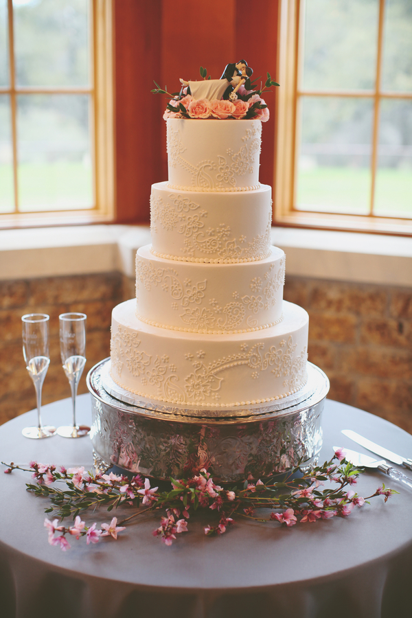 Four-tiered white wedding cake showcasing elegant wedding cake styles, adorned with delicate icing patterns and pink flowers, placed on a glass stand with floral accents beside two empty champagne flutes.