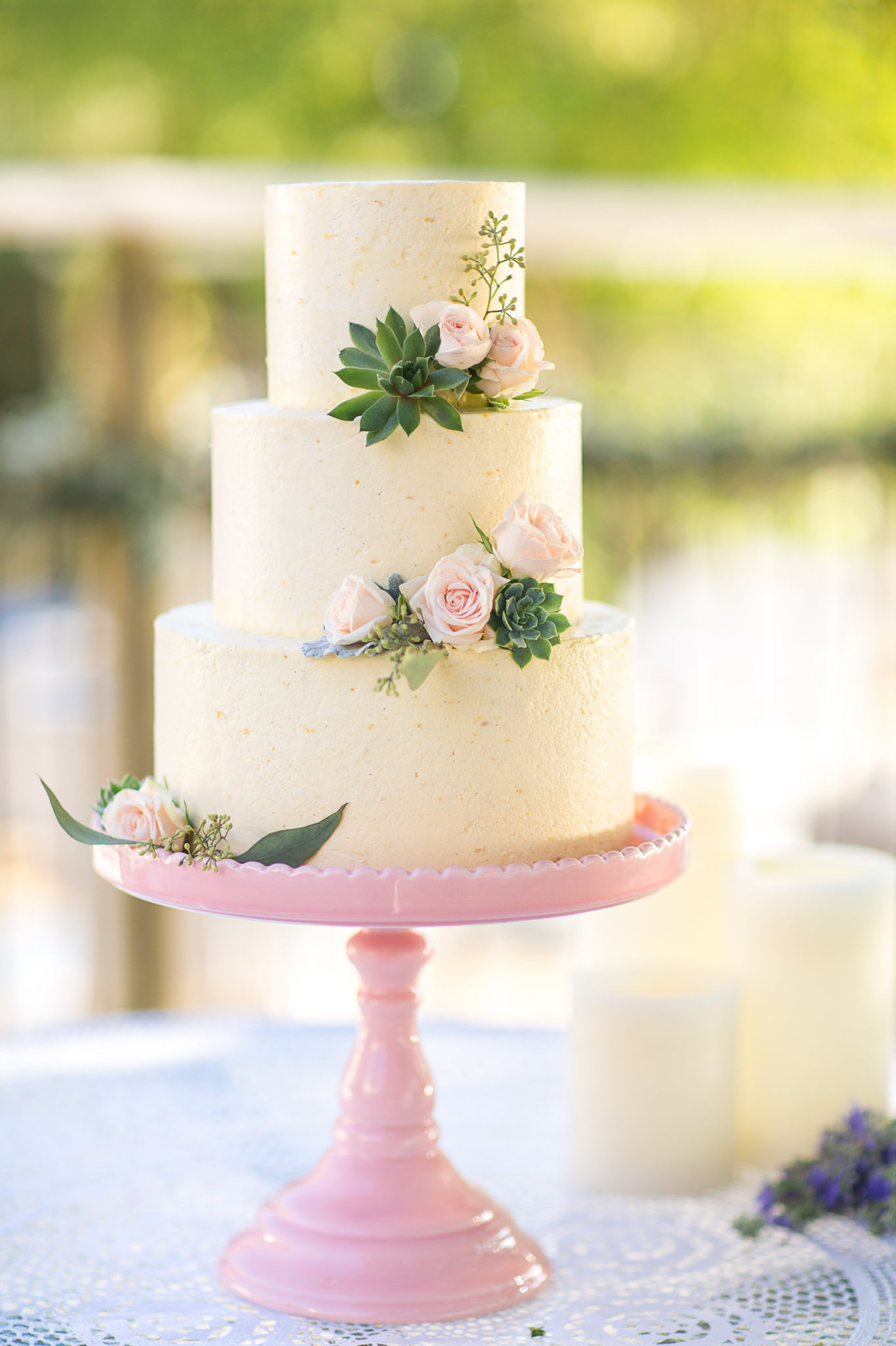 Three-tiered white cake decorated with small pink roses and green succulents, displayed on a pink cake stand outdoors—an elegant example of classic wedding cake styles.