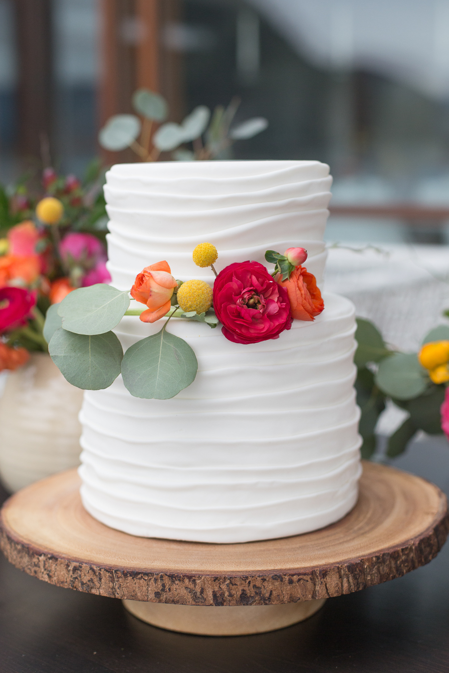 A two-tiered white cake with horizontal textured frosting—showcasing wedding cake styles—decorated with red, orange, and yellow flowers and green leaves, displayed on a wooden cake stand.
