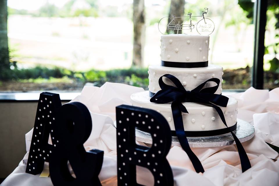 A three-tiered white cake with a black ribbon, inspired by classic wedding cake styles, sits on a table next to large black letters "A" and "B" with an ampersand, in front of a window.
