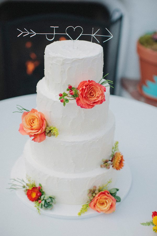 Four-tiered white wedding cake, inspired by elegant wedding cake styles, adorned with orange and red flowers and topped with a wire arrow and the initials "J" and "K" linked by a heart.