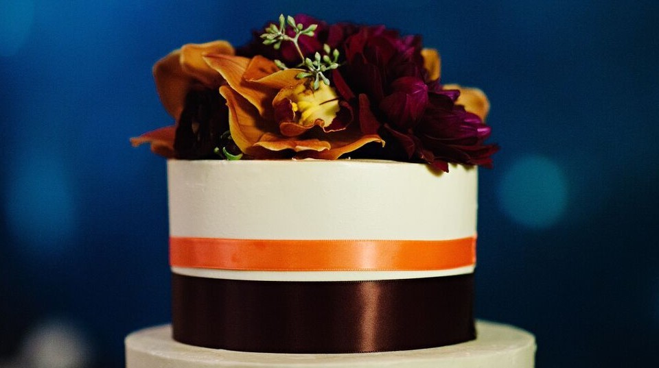 A round white cake featuring wedding cake styles with orange and dark brown ribbons, topped with orange and burgundy flowers, set against a blurred blue background.