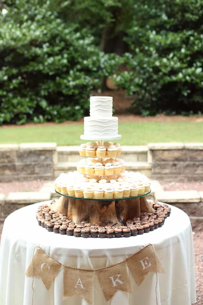 A tiered wedding cake with white frosting, showcasing elegant wedding cake styles, is displayed on a stand and surrounded by layers of vanilla and chocolate cupcakes on a round table outdoors.